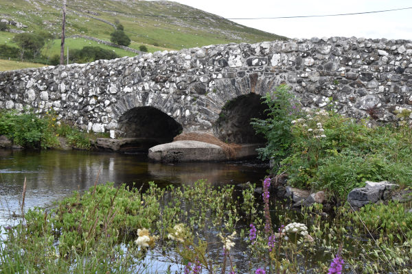 Eine alte Steinbrücke führt über einen langsam fließenden Fluss. Blumen und Gras sind im Vordergrund des Bildes sichtbar. (c) Menges