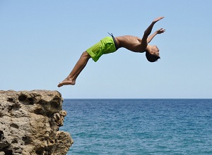 Junge in Badehose springt rückwärts von einem hohen Felsen ins Meer, Foto Credits: clicjeroendits / pixabay.com