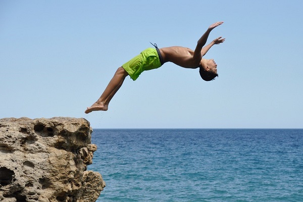 Junge in Badehose springt rückwärts von einem hohen Felsen ins Meer, Foto Credits: clicjeroendits / pixabay.com