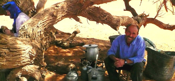 Ein nordafrikanischer Mann sitzt im Freien am Boden vor einer Menge Utensilien. Er lächelt.
