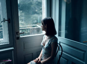 Eine Frau sitzt auf einem Stuhl und schaut durch die Glastür nach außen. Bild von Masha Raymer, Pexels.